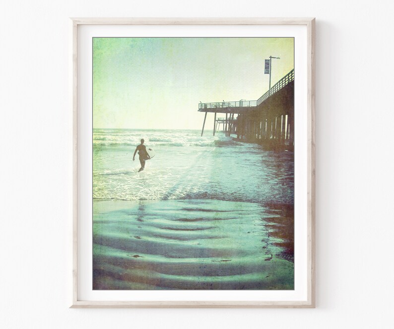 Photograph of a surfer walking out to the ocean next to a wooden pier at Pismo beach California, with a vintage style. Fine art photography print.