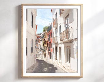 Puede incluir: Una impresión artística enmarcada que representa una calle europea estrecha y soleada. La imagen muestra edificios blancos con balcones, una fachada roja y un camino empedrado que conduce a la distancia bajo un cielo azul claro y brillante.