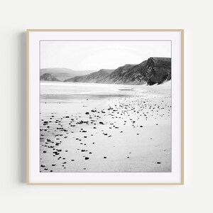 Puede incluir: Una fotograf&iacute;a enmarcada en blanco y negro de un paisaje costero que muestra una playa de arena salpicada de peque&ntilde;as piedras oscuras, que conduce hacia una orilla con colinas y acantilados a lo lejos bajo un cielo brillante y nublado.