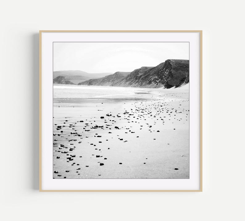 Puede incluir: Una fotograf&iacute;a enmarcada en blanco y negro de un paisaje costero que muestra una playa de arena salpicada de peque&ntilde;as piedras oscuras, que conduce hacia una orilla con colinas y acantilados a lo lejos bajo un cielo brillante y nublado.