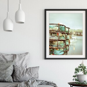 May include: Framed art print featuring a coastal scene. The image showcases a stack of weathered crab traps with turquoise rope, set against a backdrop of a calm harbor and boats. The artwork is framed in a black border.