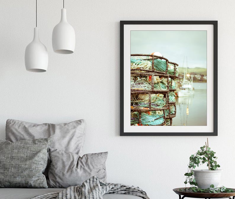May include: Framed art print featuring a coastal scene. The image showcases a stack of weathered crab traps with turquoise rope, set against a backdrop of a calm harbor and boats. The artwork is framed in a black border.