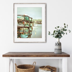 May include: A framed print of a coastal scene featuring a stack of crab traps with turquoise rope and buoys. A white boat is visible in the background, with a muted color palette. The print is displayed on a wooden console table with woven baskets.