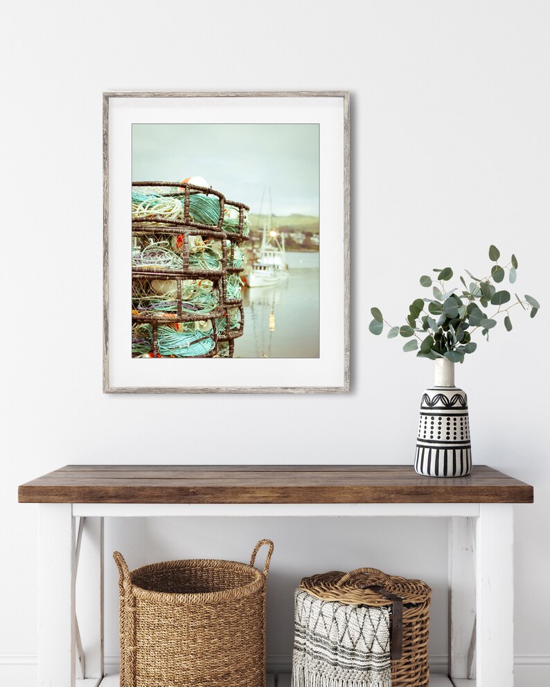May include: A framed print of a coastal scene featuring a stack of crab traps with turquoise rope and buoys. A white boat is visible in the background, with a muted color palette. The print is displayed on a wooden console table with woven baskets.