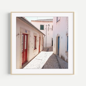 May include: A framed print of a narrow cobblestone street in a European town. White buildings with red doors and window frames line the street. The image is bathed in sunlight, creating strong shadows. A blue door is visible in the distance.