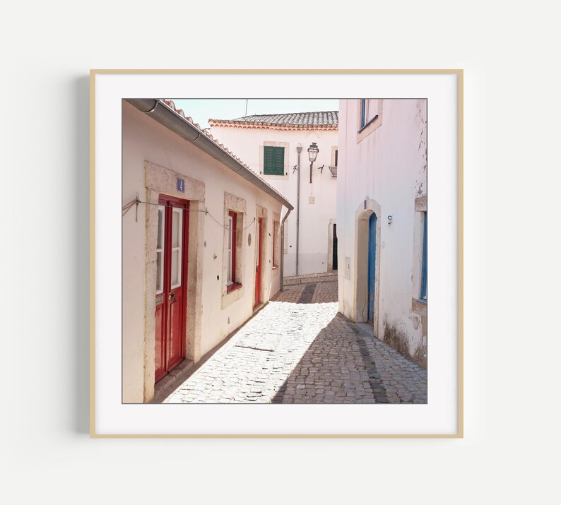 May include: A framed print of a narrow cobblestone street in a European town. White buildings with red doors and window frames line the street. The image is bathed in sunlight, creating strong shadows. A blue door is visible in the distance.