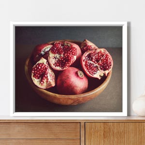 May include: A framed wall art print featuring a wooden bowl filled with several pomegranates, including whole fruits and halved sections displaying deep red seeds, set against a dark, moody background with a white frame border.