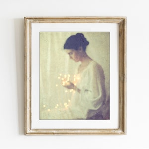 May include: A framed art print featuring a soft, vintage-style photograph. The image shows a person in a white dress holding glowing fairy lights. The frame is a light, distressed gold color, adding to the artwork's ethereal quality.