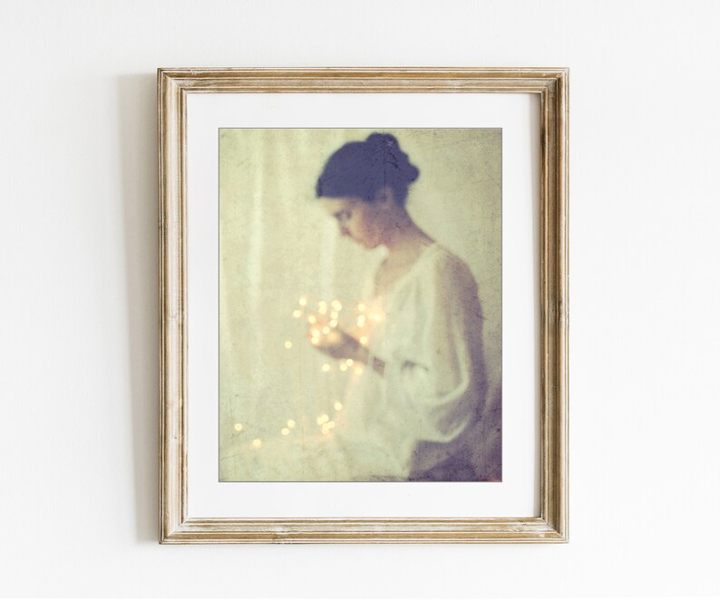 May include: A framed art print featuring a soft, vintage-style photograph. The image shows a person in a white dress holding glowing fairy lights. The frame is a light, distressed gold color, adding to the artwork's ethereal quality.