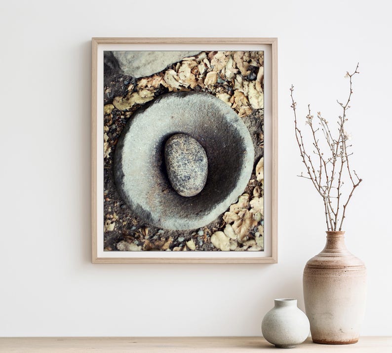 May include: A framed wall art print featuring a top-down view of a smooth, circular grey stone mortar with a smaller, speckled grey stone pestle resting inside, surrounded by a scattering of dried, light brown autumn leaves on the ground.