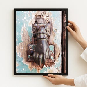 May include: A framed wall art print featuring a weathered, teal-painted wooden door with a dark, tarnished metal door knocker shaped like a human hand. The paint on the door is heavily chipped, revealing the natural wood grain underneath the surface.