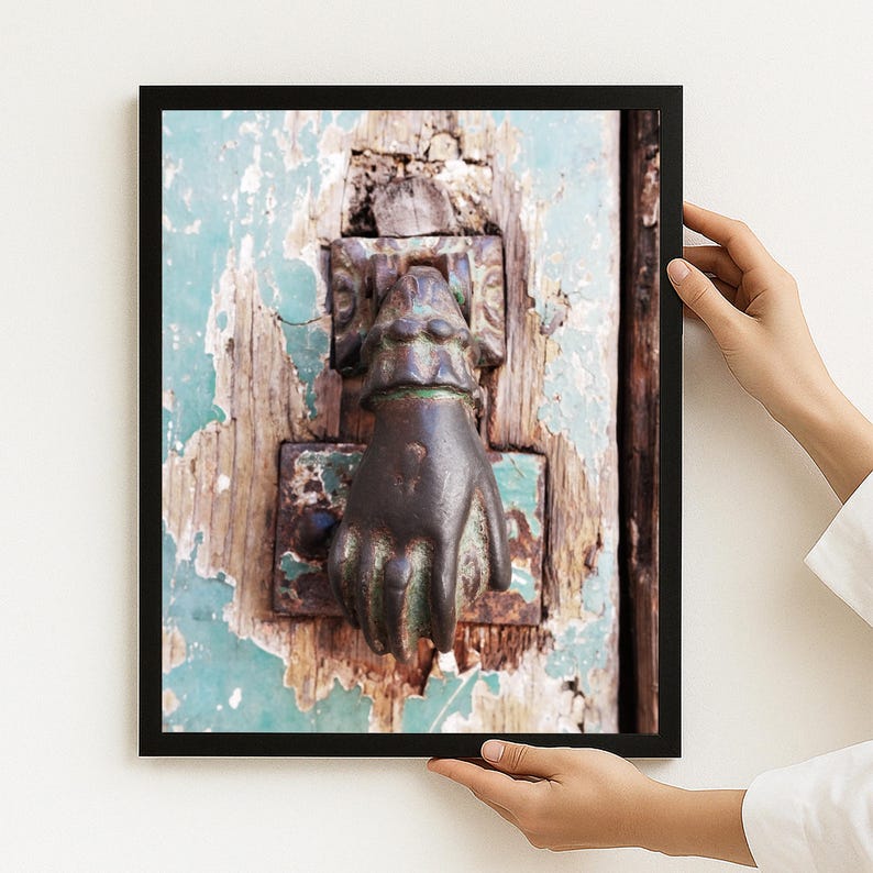 May include: A framed wall art print featuring a weathered, teal-painted wooden door with a dark, tarnished metal door knocker shaped like a human hand. The paint on the door is heavily chipped, revealing the natural wood grain underneath the surface.