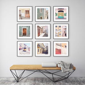 May include: Nine framed art prints featuring architectural scenes. The prints showcase colorful doors, buildings, and streets. The artwork is displayed above a wooden bench with a black metal base, creating a gallery wall aesthetic.