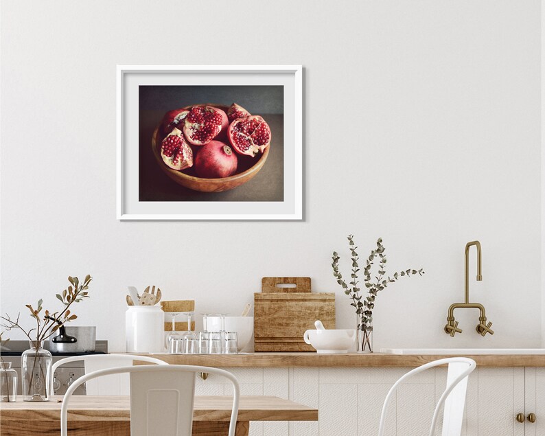 May include: A framed photograph of a wooden bowl filled with red pomegranate fruit. The bowl is on a brown surface. The background is a dark gray color.