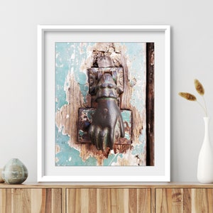 May include: A framed wall art print featuring a dark metal hand-shaped door knocker mounted on a weathered, teal-painted wooden door with visible wood grain and peeling paint, displayed in a white frame on a wooden surface with decorative vases.