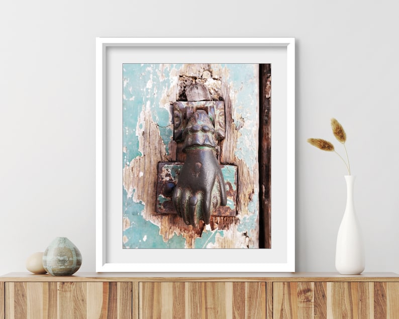 May include: A framed wall art print featuring a dark metal hand-shaped door knocker mounted on a weathered, teal-painted wooden door with visible wood grain and peeling paint, displayed in a white frame on a wooden surface with decorative vases.