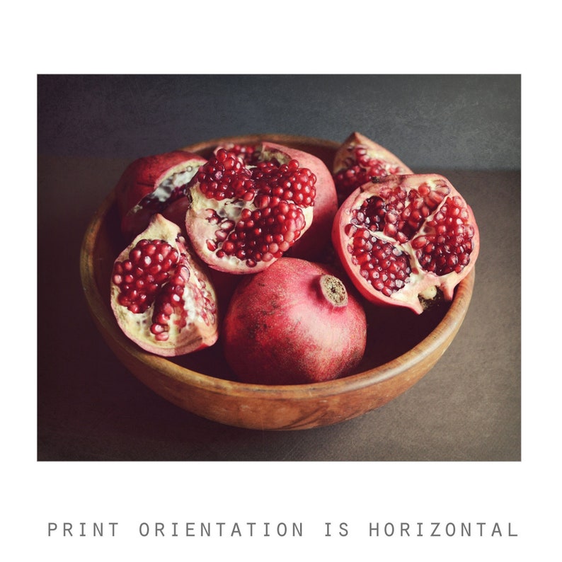 May include: A wooden bowl filled with several pomegranates, including one whole fruit and multiple pieces showing the deep red, glistening arils inside. The fruit has a textured, reddish-pink rind and is arranged in a rustic, dark-toned setting.