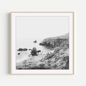 Puede incluir: Una impresi&oacute;n de arte mural cuadrada en blanco y negro que representa un paisaje costero con un acantilado escarpado sobre una playa de arena, formaciones rocosas dispersas en el agua y un horizonte brumoso, todo en un marco de madera clara.