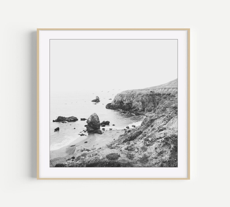 Puede incluir: Una impresi&oacute;n de arte mural cuadrada en blanco y negro que representa un paisaje costero con un acantilado escarpado sobre una playa de arena, formaciones rocosas dispersas en el agua y un horizonte brumoso, todo en un marco de madera clara.