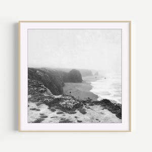 Puede incluir: Una fotograf&iacute;a cuadrada en blanco y negro enmarcada que representa un acantilado costero escarpado con vistas a una playa de arena y las olas del oc&eacute;ano. La escena est&aacute; envuelta en una espesa niebla, con vegetaci&oacute;n baja en primer plano.