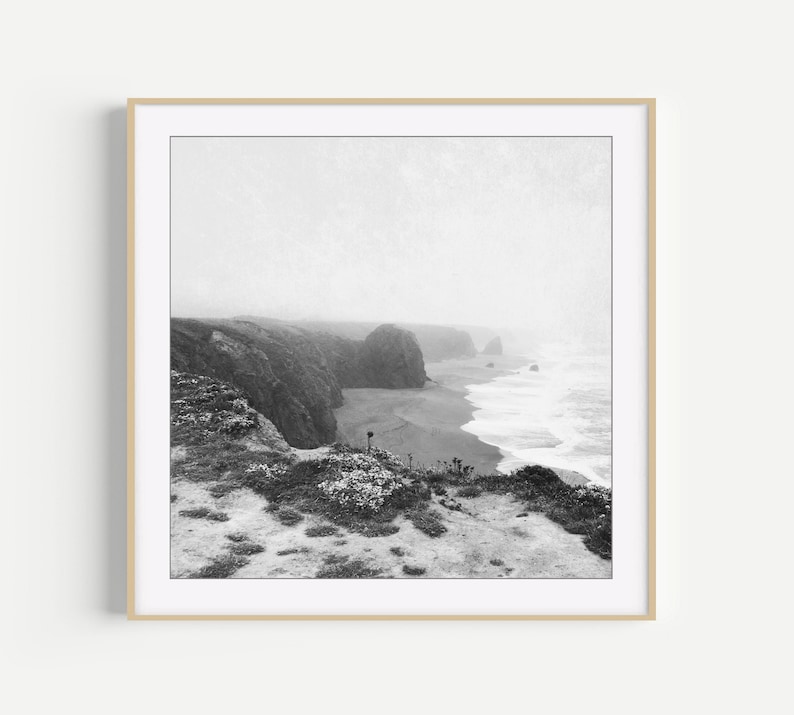 Puede incluir: Una fotograf&iacute;a cuadrada en blanco y negro enmarcada que representa un acantilado costero escarpado con vistas a una playa de arena y las olas del oc&eacute;ano. La escena est&aacute; envuelta en una espesa niebla, con vegetaci&oacute;n baja en primer plano.
