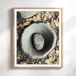 May include: A framed wall art print featuring a top-down view of a circular, weathered grey stone mortar with a smooth, speckled grey stone pestle resting in the centre, surrounded by a scattering of dry, light brown fallen autumn leaves.