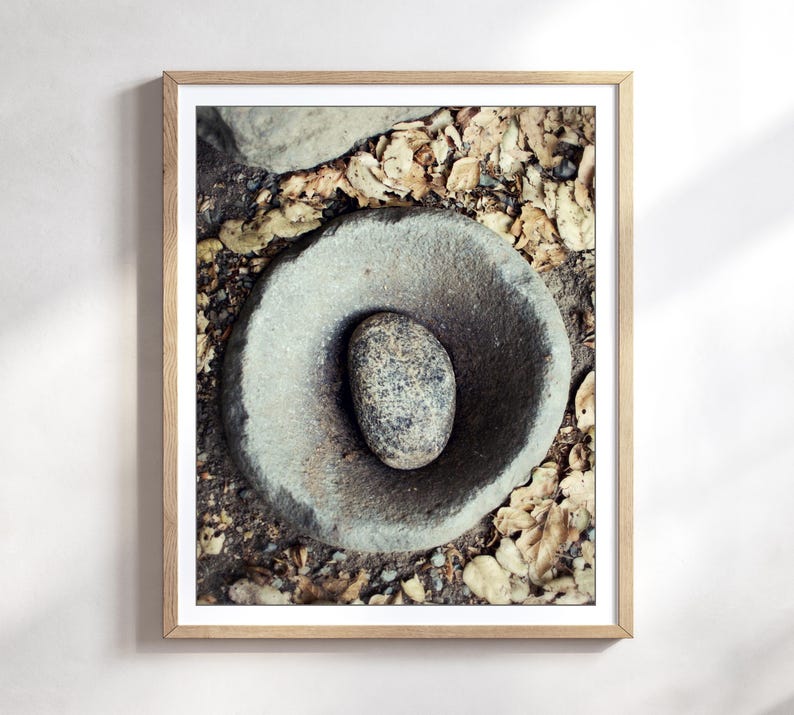 May include: A framed wall art print featuring a top-down view of a circular, weathered grey stone mortar with a smooth, speckled grey stone pestle resting in the centre, surrounded by a scattering of dry, light brown fallen autumn leaves.
