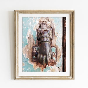 May include: A framed wall art print featuring a weathered, dark metal door knocker shaped like a hand, mounted on a rustic, distressed wooden door with peeling teal paint. The frame is a light, textured gold color with a white mat border.