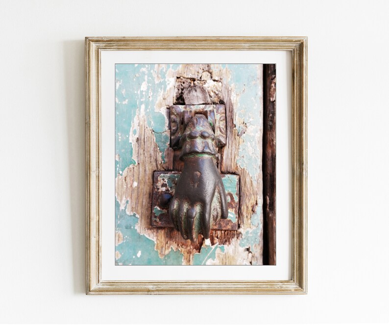May include: A framed wall art print featuring a weathered, dark metal door knocker shaped like a hand, mounted on a rustic, distressed wooden door with peeling teal paint. The frame is a light, textured gold color with a white mat border.