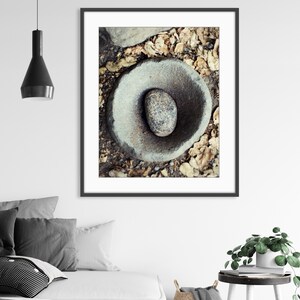 May include: A framed wall art print featuring a top-down view of a circular stone mortar and pestle set. The grey, weathered stone bowl contains a smooth, speckled grey stone pestle, all resting on a ground surface covered with dry brown leaves.