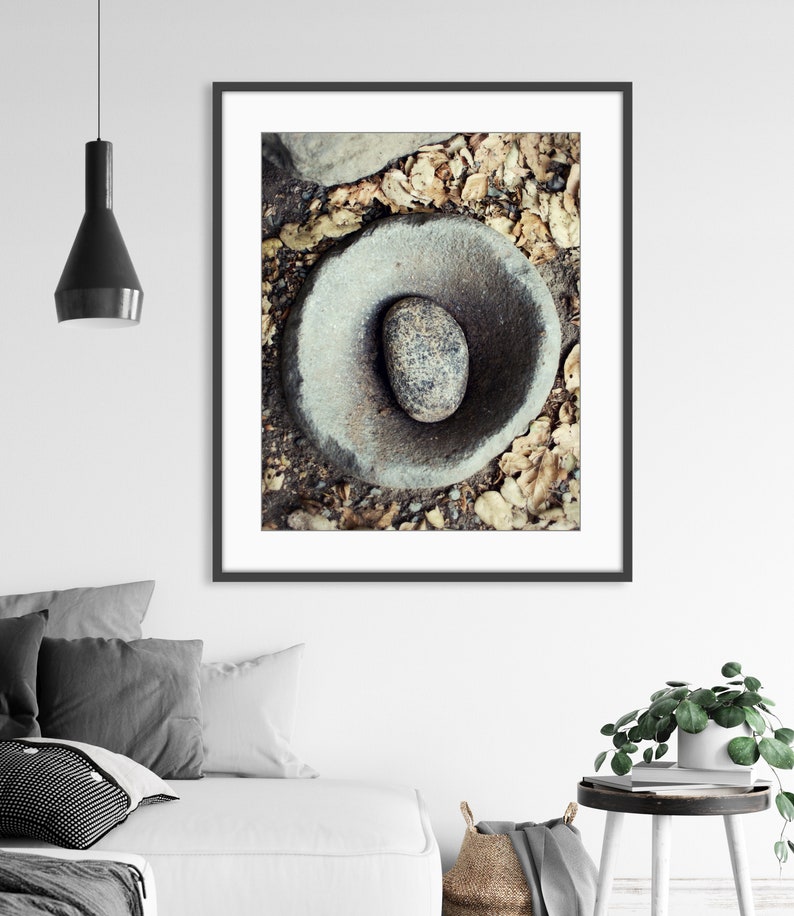 May include: A framed wall art print featuring a top-down view of a circular stone mortar and pestle set. The grey, weathered stone bowl contains a smooth, speckled grey stone pestle, all resting on a ground surface covered with dry brown leaves.