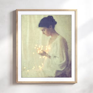 May include: A framed art print featuring a soft, vintage-style image of a person holding glowing lights. The person is wearing a white top, and the overall color palette is muted with warm tones.