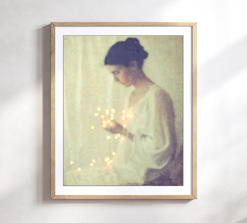 May include: A framed art print featuring a soft, vintage-style image of a person holding glowing lights. The person is wearing a white top, and the overall color palette is muted with warm tones.