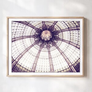 May include: Framed print of a detailed architectural ceiling. The image showcases a symmetrical design with glass panels and intricate metalwork, in shades of purple and white. The artwork is displayed in a light-colored wooden frame.