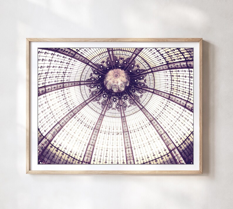 May include: Framed print of a detailed architectural ceiling. The image showcases a symmetrical design with glass panels and intricate metalwork, in shades of purple and white. The artwork is displayed in a light-colored wooden frame.