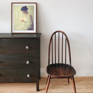 May include: A dark brown wooden dresser with three drawers and decorative knobs. A framed picture hangs above the dresser. A wooden chair with a curved back and spindle supports sits nearby. The room has light-colored wood flooring.