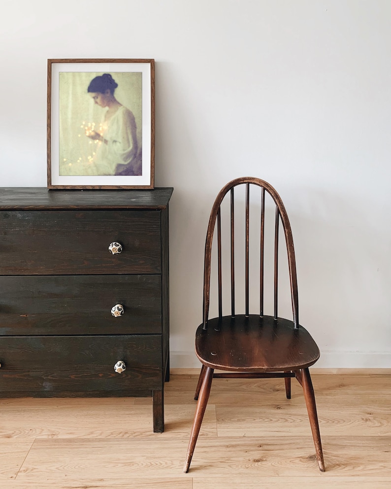 May include: A dark brown wooden dresser with three drawers and decorative knobs. A framed picture hangs above the dresser. A wooden chair with a curved back and spindle supports sits nearby. The room has light-colored wood flooring.