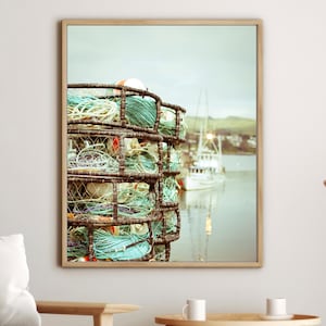 May include: A framed photograph of a fishing scene. A stack of crab traps filled with colorful ropes and buoys is in the foreground. A boat is docked in the background on a cloudy day. The image has a vintage aesthetic.