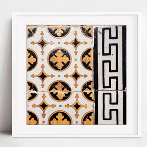 May include: A framed print of a close-up of a vintage tile pattern. The tile is white with black and gold geometric designs. The tile is cracked in the center.