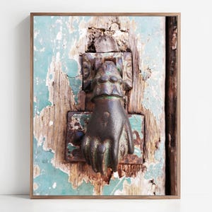 May include: A dark bronze-colored metal door knocker shaped like a hand, mounted on a weathered wooden door with peeling teal paint. The knocker features a cuff detail at the wrist and is attached to a rectangular metal plate with visible rust.