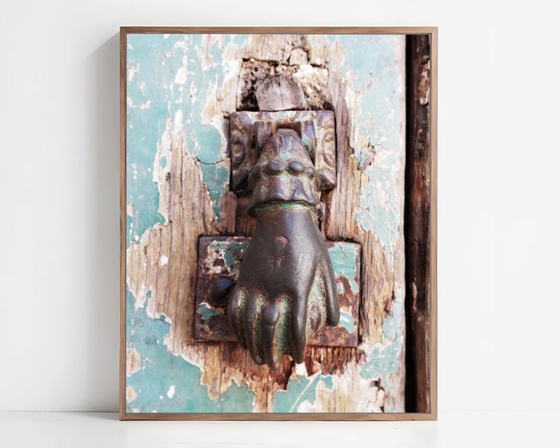 May include: A dark bronze-colored metal door knocker shaped like a hand, mounted on a weathered wooden door with peeling teal paint. The knocker features a cuff detail at the wrist and is attached to a rectangular metal plate with visible rust.