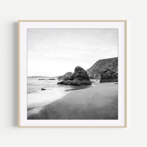 Puede incluir: Una fotograf&iacute;a cuadrada en blanco y negro que muestra una playa de arena con grandes formaciones rocosas irregulares que emergen de la orilla. Las olas del mar ba&ntilde;an suavemente la arena oscura, mientras un acantilado rocoso se alza al fondo.