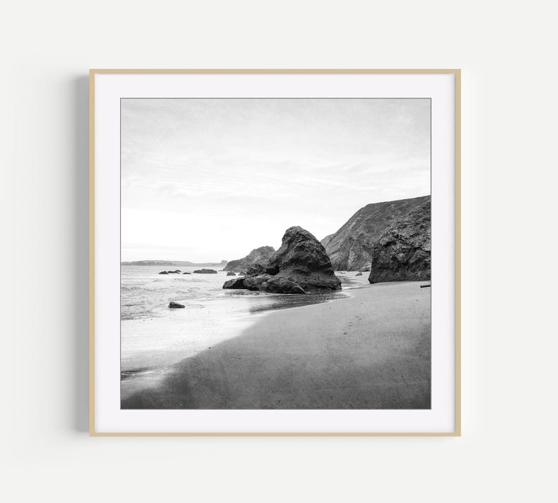 Puede incluir: Una fotograf&iacute;a cuadrada en blanco y negro que muestra una playa de arena con grandes formaciones rocosas irregulares que emergen de la orilla. Las olas del mar ba&ntilde;an suavemente la arena oscura, mientras un acantilado rocoso se alza al fondo.