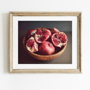 May include: A framed wall art print featuring a wooden bowl filled with several whole and halved pomegranates. The fruit displays deep red skins and clusters of bright red arils. The frame is a light-toned wood with a white mat border.