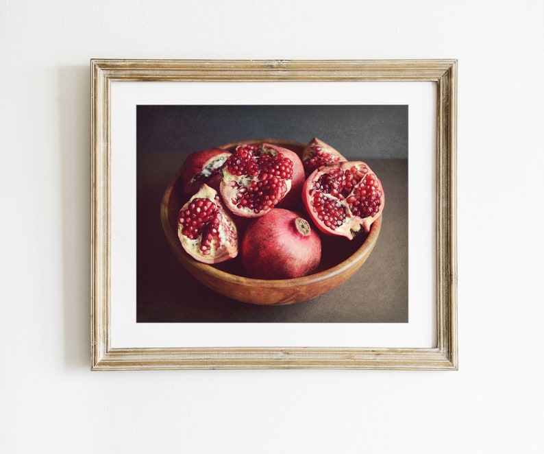 May include: A framed wall art print featuring a wooden bowl filled with several whole and halved pomegranates. The fruit displays deep red skins and clusters of bright red arils. The frame is a light-toned wood with a white mat border.