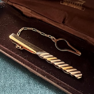 Könnte beinhalten: Eine goldfarbene Krawattenklammer mit einem strukturierten, gedrehten Design und einer passenden Kette. Die Klammer wird in einem braunen Etui mit den Initialen "MP" und einem dekorativen Emblem präsentiert.