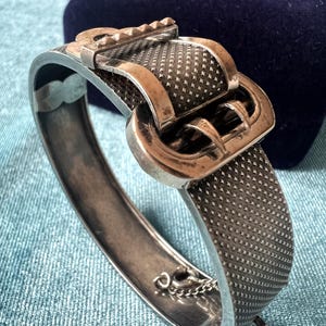 May include: A silver-toned metal hinged bangle bracelet featuring a textured, cross-hatched surface pattern. The bracelet is designed with a prominent buckle-style clasp mechanism on the front, complete with a metal pin and a safety chain attachment.