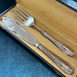 May include: A silver-toned fish serving set consisting of a wide, flat knife and a four-tined fork, both featuring ornate, embossed scrollwork patterns on the handles and blades, presented in a rectangular, dark-colored hinged storage case.