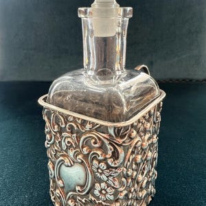 Op de afbeelding: Een karaf van helder glas met een bijpassende glazen stop, geplaatst in een decoratieve zilverkleurige metalen houder. De metalen houder is voorzien van gedetailleerde reliëfpatronen met bloemen en krullen, inclusief een glad hartvormig schild.