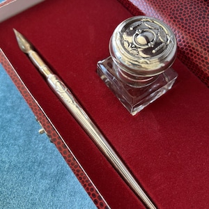 Peut inclure: Un porte-plume en métal argenté et un encrier en verre carré avec un couvercle décoratif en métal argenté, présentés dans un coffret rectangulaire doublé de velours rouge. Le porte-plume possède une plume pointue et un manche effilé gravé.